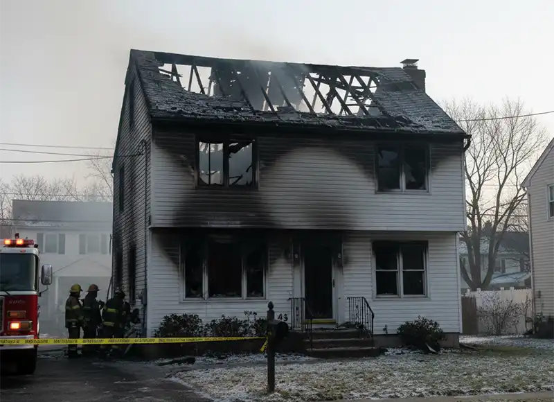 Firefighters assessing smoke-damaged two-story white home with burned-out roof